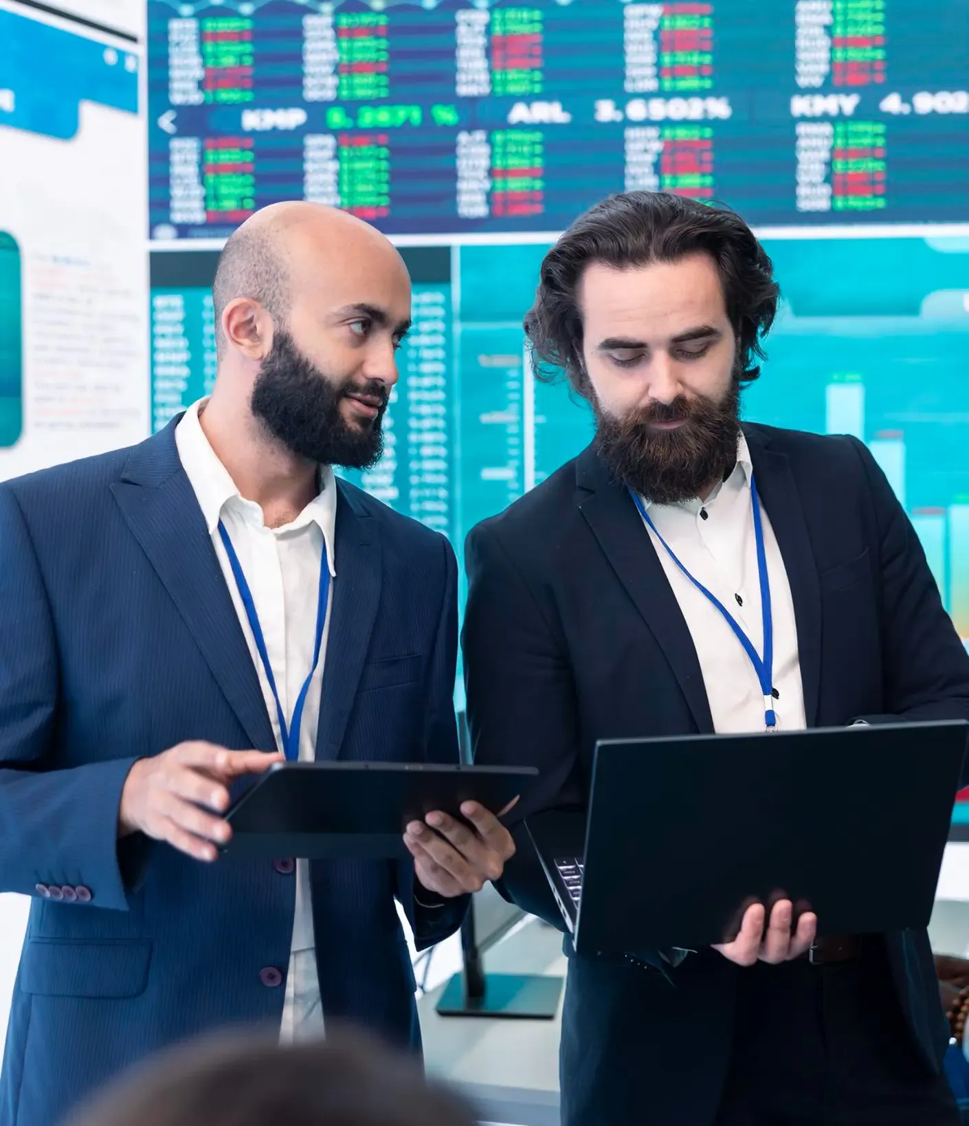 Two businessmen in suits discuss data on a tablet and laptop, with stock market screens in the background.
