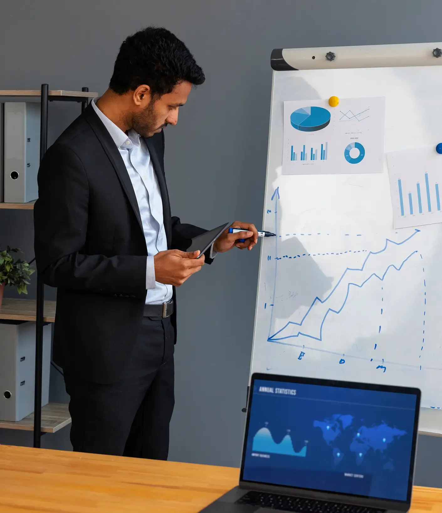 Man in suit presents data on a flipchart with graphs, holding a tablet. Laptop shows annual statistics on the table.