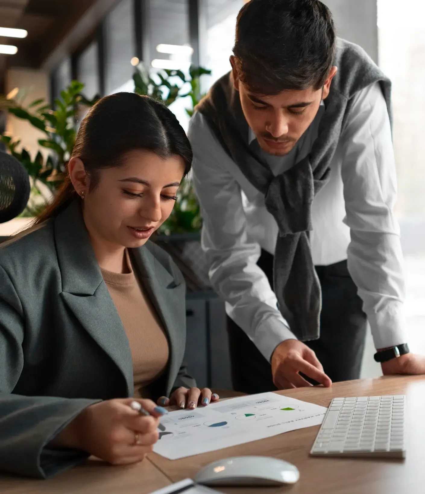Two colleagues collaborate on a project, reviewing charts at a desk in a modern office with plants and natural light.
